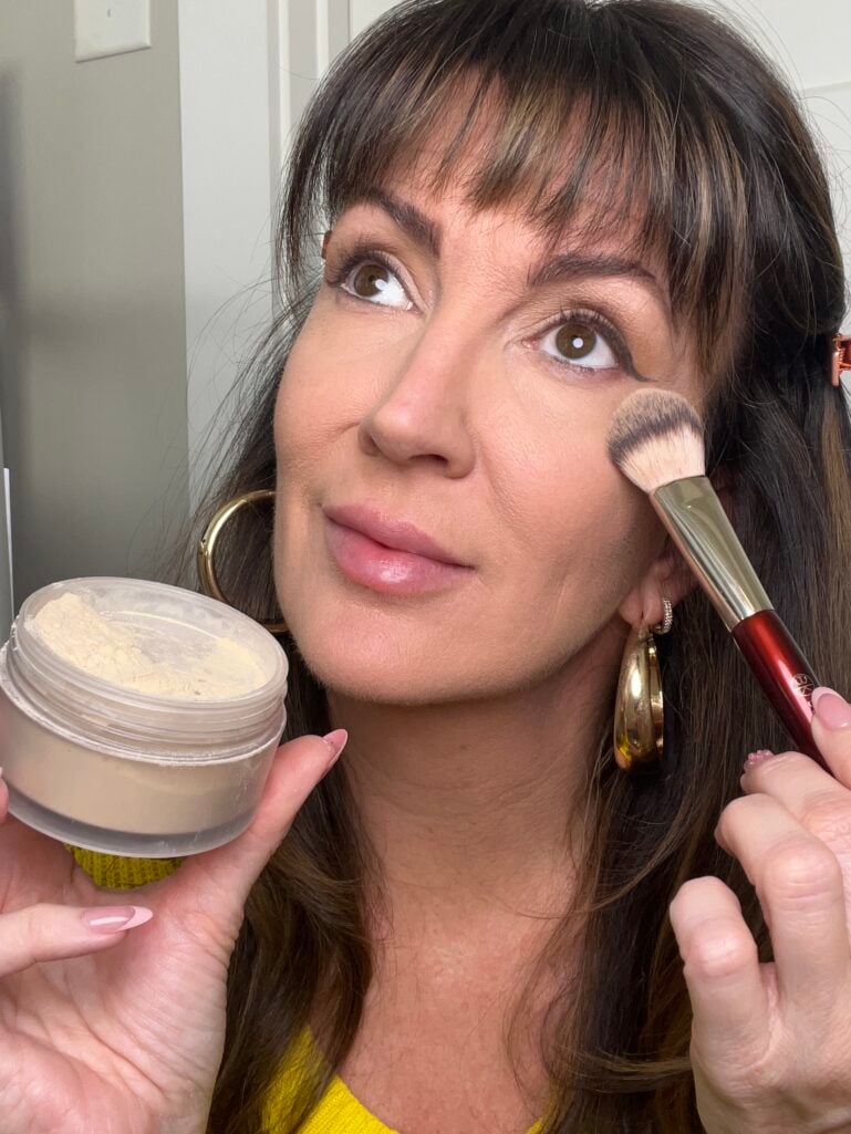 Woman applying Laura Mercier Ultra Blur loose powder to cheek with BK Beauty brush while holding open powder jar showing translucent yellow-tinted powder, wearing yellow sweater, gold puffy hoop earrings, copper hair clips, pink French tip nails, looking upward
