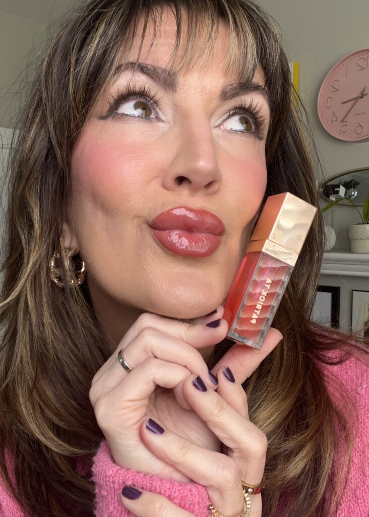 Close-up of woman looking upward showing finished Valentine's Day lip look with glossy plumped rose lips, holding Patrick Ta Major Plumping lip gloss in shade Full Syringe with red-orange gradient packaging, rosy flushed cheeks, soft smokey eyes, gold hoop earrings, pink clock visible in background