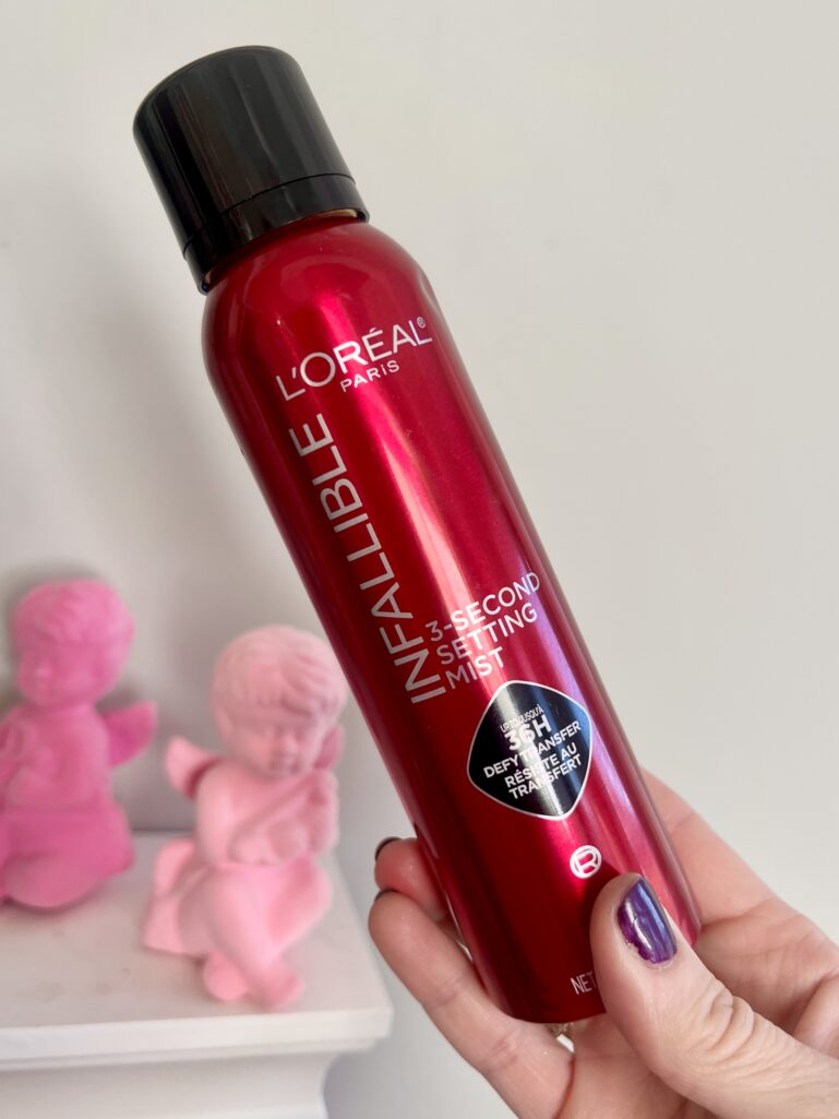 Hand holding red L'Oreal Infallible 3-Second Setting Mist spray bottle with black cap, pink flocked cupid figurines in background on white shelf, dark purple nail polish visible