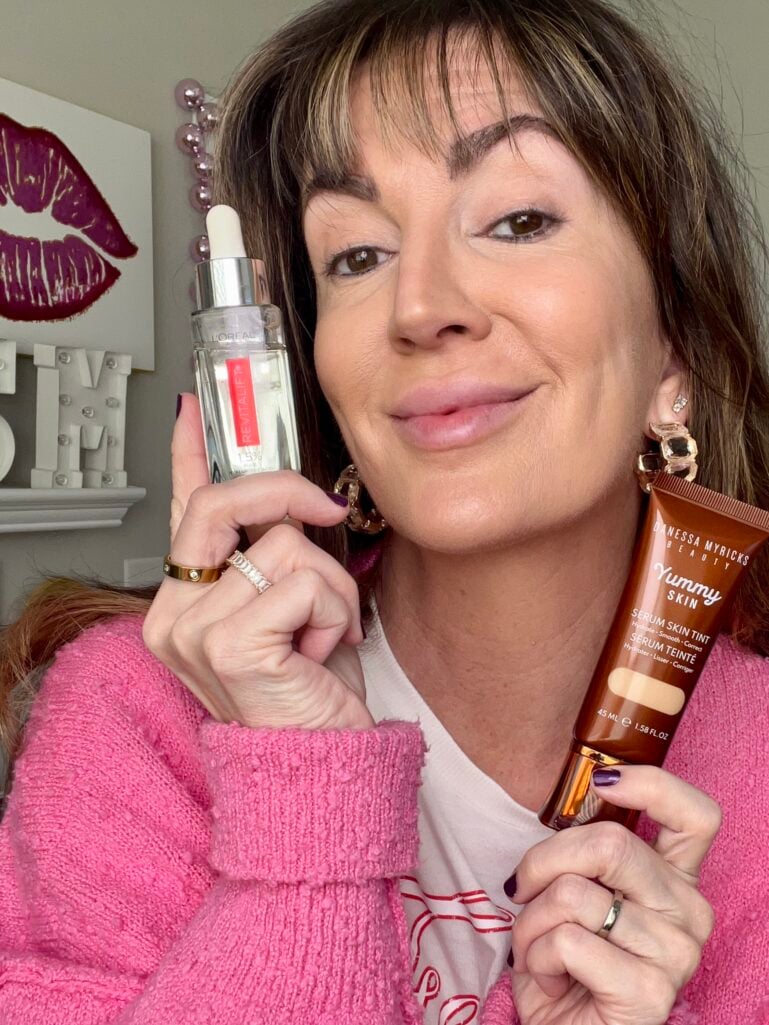 Woman with bare face holding L'Oreal Revitalift Pure Hyaluronic Acid serum in one hand and Danessa Myricks Yummy Skin Serum Tint tube in other, wearing pink cardigan, gold crystal hoop earrings, dark purple nail polish, lips art print and pink beaded mirror frame visible in background