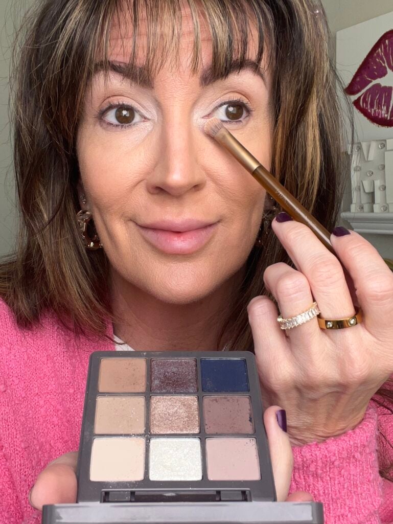 Woman applying eyeshadow with gold brush while holding open Anastasia Beverly Hills Haze 9-shade palette showing neutral mattes, shimmers, and navy shades, wearing pink cardigan, gold hoop earrings, gold Cartier-style ring stack, dark purple nail polish, lips art visible in background