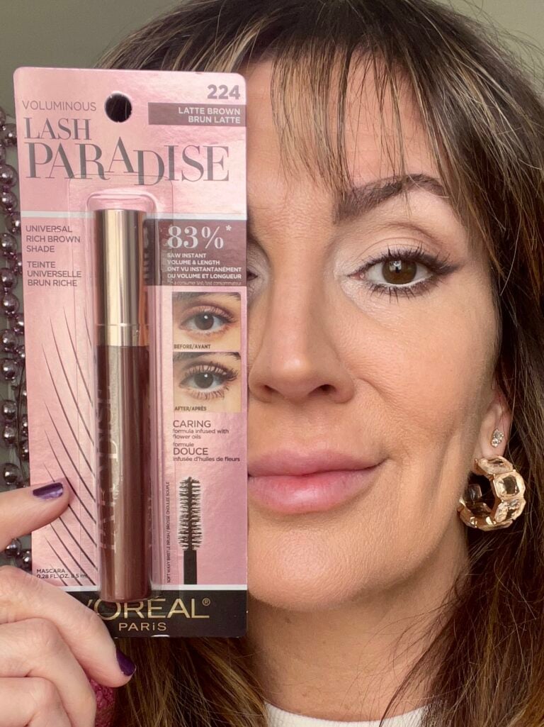 Close-up of woman holding L'Oreal Voluminous Lash Paradise mascara package in shade 224 Latte Brown next to face, showing soft eye makeup with brown mascara on lashes, gold chunky crystal hoop earrings, pink beaded mirror frame in background