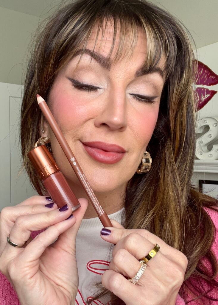 Woman with eyes closed showing completed smokey eye with brown cat eye liner, holding Clarins lip water stain in nude brown shade and Morphe lip pencil in shade Whipped, wearing pink cardigan, gold crystal hoop earrings, gold Cartier-style ring, dark purple nails, rose-stained lips