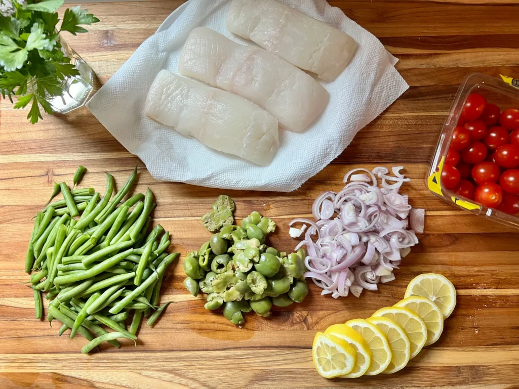 Overhead view of ingredients on a wooden cutting board: three halibut filets on paper towels, halved green beans, smashed green olives, thinly sliced shallots, lemon slices, cherry tomatoes, and fresh herbs in a glass