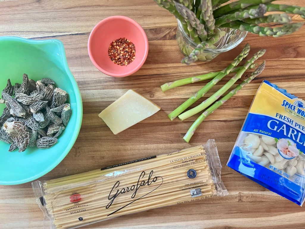 Overhead view of ingredients on a wooden cutting board: dried morel mushrooms in a green bowl, Garofalo bucatini, fresh asparagus, Parmesan wedge, crushed red pepper in a pink bowl, and a bag of fresh peeled garlic