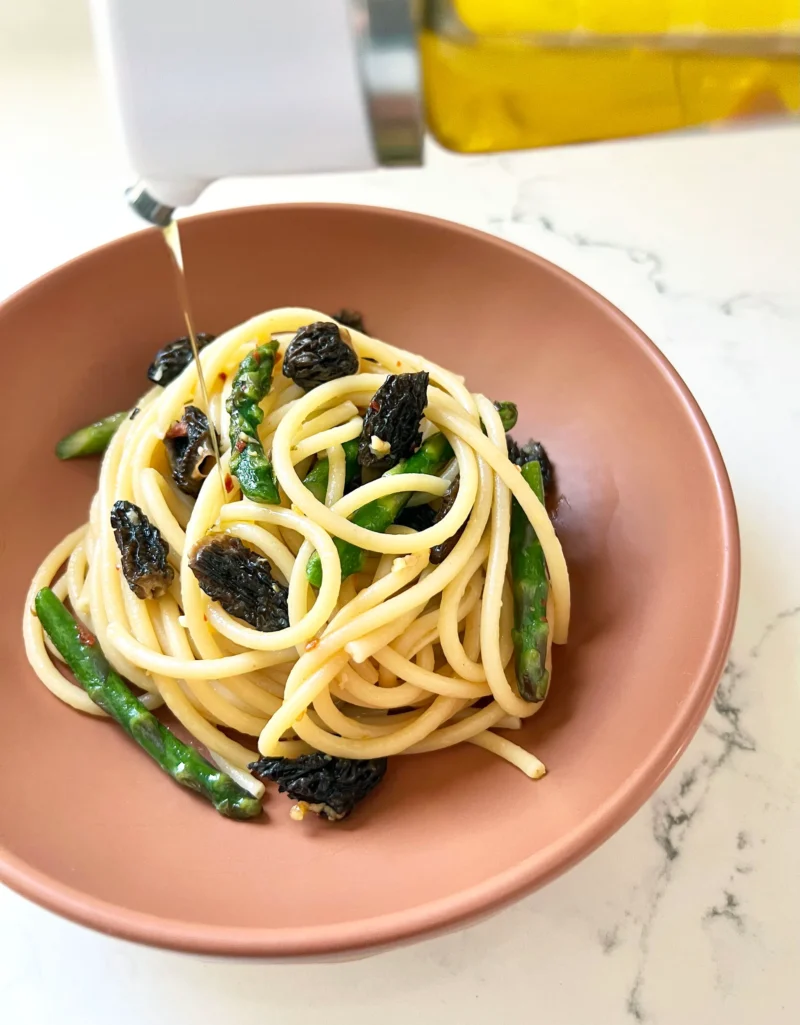 Plated bucatini with asparagus and morels in a terracotta bowl with a drizzle of olive oil being added from a bottle in the background