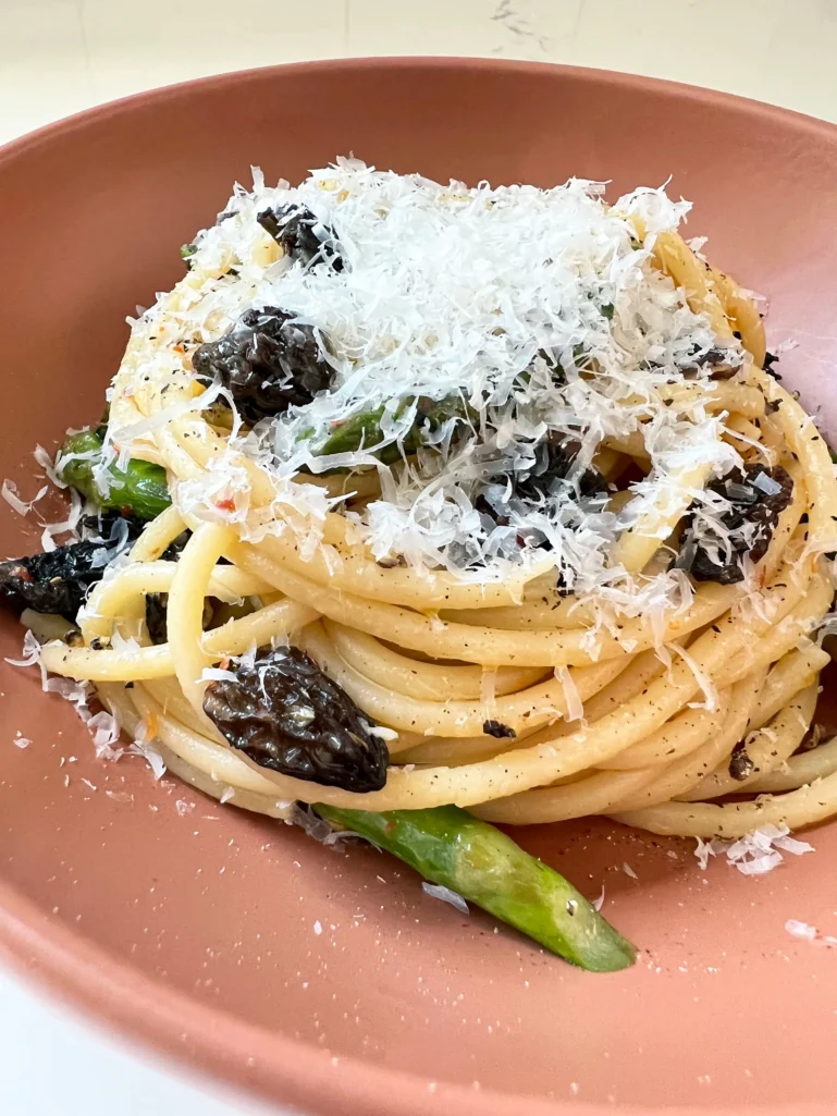 Close-up of finished bucatini with asparagus and morels topped with freshly grated Parmesan and cracked black pepper in a terracotta bowl