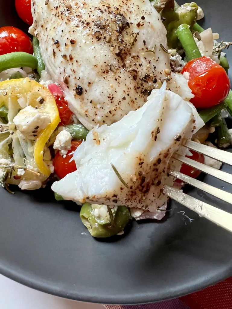 Close-up of flaky baked halibut with cracked black pepper being pulled apart with a gold fork, showing the tender white interior, over roasted vegetables with olives, tomatoes, and feta