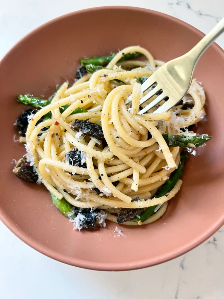 Overhead view of bucatini with asparagus and morels twirled around a gold fork in a terracotta bowl with grated Parmesan