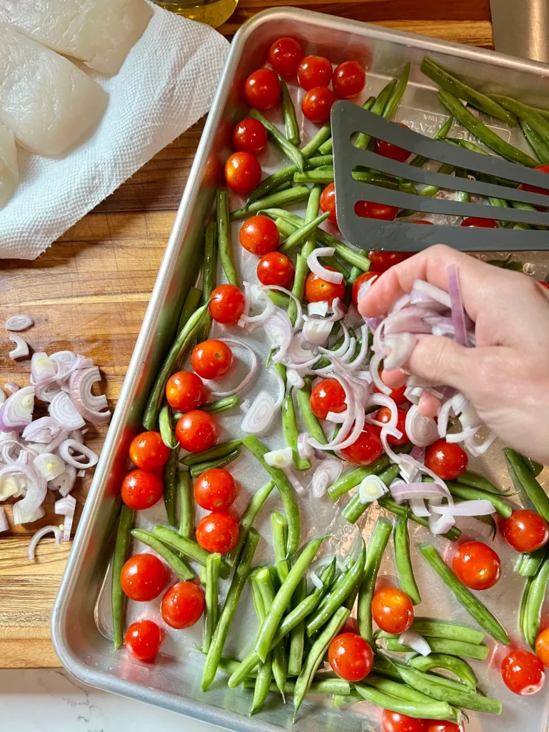 Hand scattering sliced shallots over green beans and cherry tomatoes on an oiled sheet pan with halibut filets on paper towels nearby