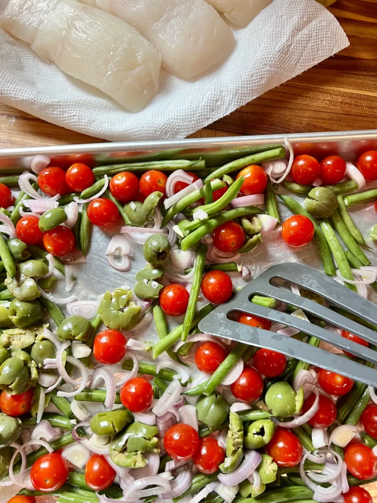 Sheet pan with green beans, cherry tomatoes, shallots, and smashed green olives tossed together with a spatula, halibut filets waiting on a cutting board
