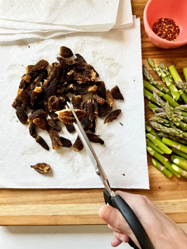 Hand using kitchen shears to cut reconstituted morel mushrooms in half on a paper towel-lined cutting board with asparagus and crushed red pepper nearby