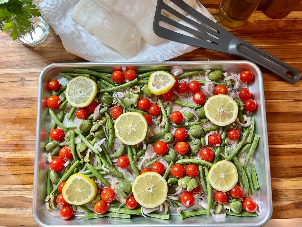 Overhead view of the sheet pan with green beans, tomatoes, shallots, olives, and lemon slices arranged on top, with halibut, fresh herbs, olive oil, and a spatula nearby