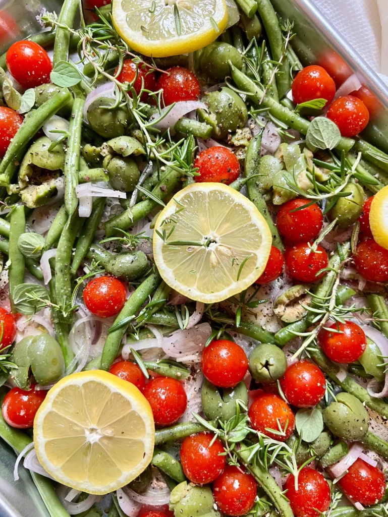 Close-up of the sheet pan with sprigs of fresh rosemary, thyme, and oregano laid over the vegetables, lemon slices, and olives