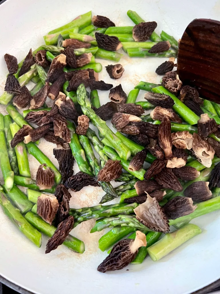 Reconstituted morel mushrooms and asparagus pieces sautéing together in a white skillet with a wooden spoon