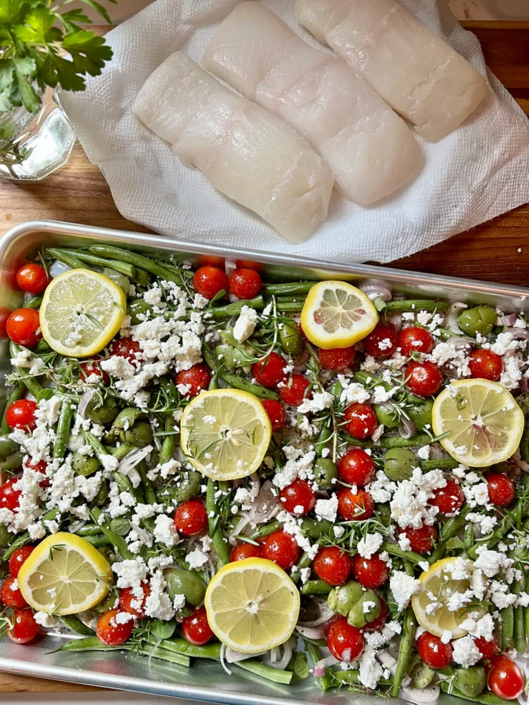 Sheet pan with crumbled feta scattered over the vegetables, herbs, and lemon slices with halibut filets on paper towels and fresh herbs in the background