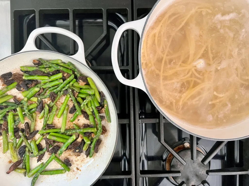 Overhead view of the stovetop with asparagus and morels in a skillet alongside a pot of bucatini boiling in salted water