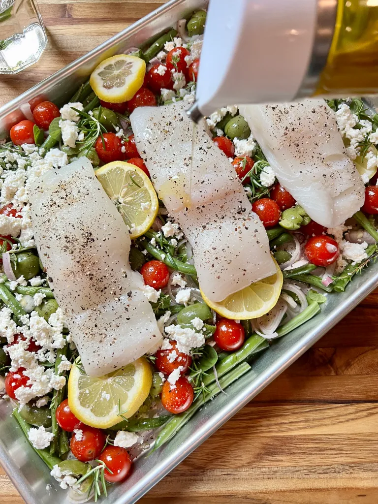 Three seasoned halibut filets placed on top of the vegetable bed on the sheet pan with olive oil being drizzled from a bottle