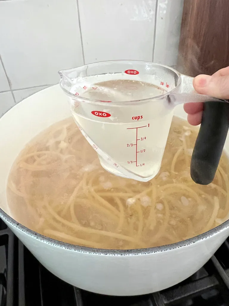 Hand holding an OXO measuring cup filled with reserved starchy pasta water over the pot of cooked bucatini