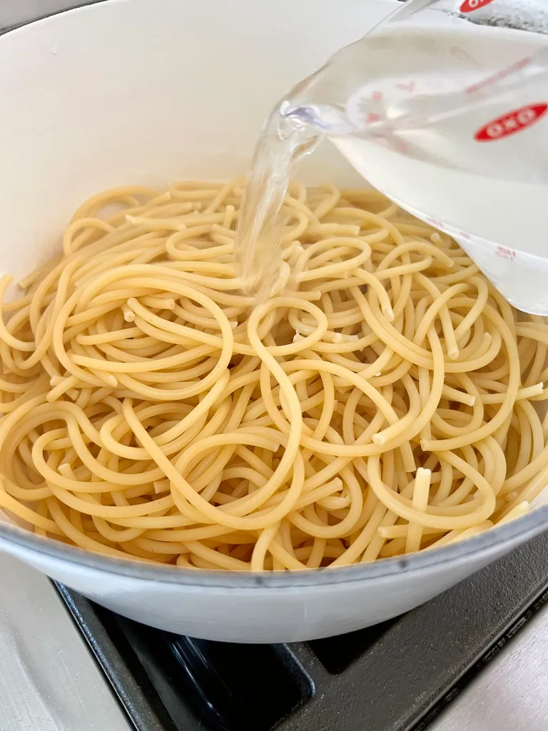 Pouring reserved pasta water from a measuring cup over drained bucatini in a white pot