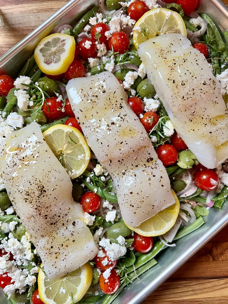 Close-up of the fully assembled sheet pan with three halibut filets seasoned with salt and pepper resting on a bed of green beans, tomatoes, olives, feta, lemon, and herbs, ready for the oven