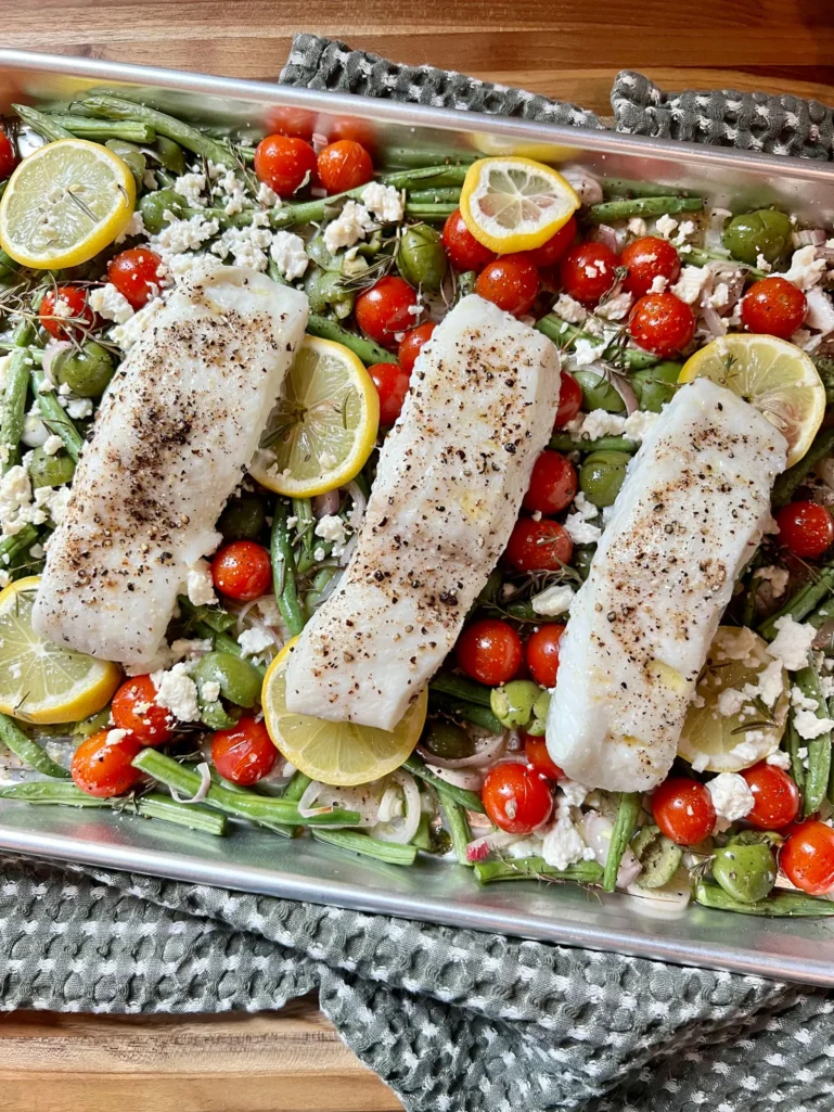 Sheet pan fresh from the oven with three perfectly cooked halibut filets on top of the roasted vegetables, resting on a gray kitchen towel
