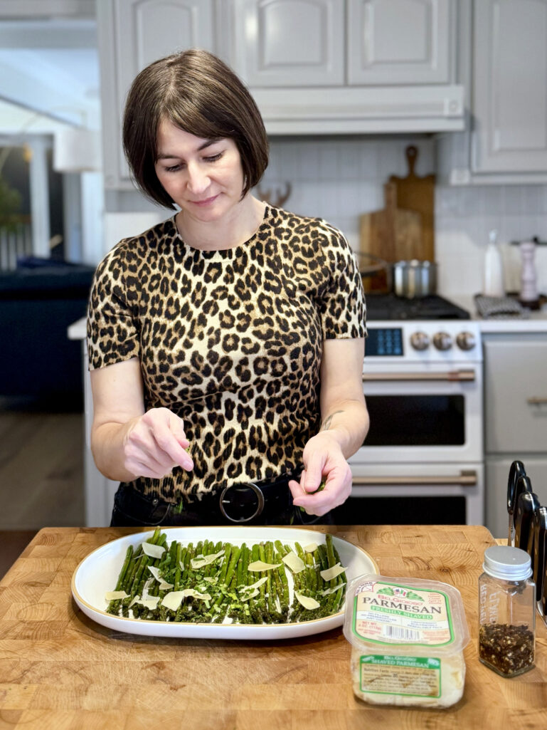 Kelly placing shaved Parmesan over a platter of roasted asparagus in the kitchen, wearing a leopard print sweater, with a container of freshly shaved Parmesan and a pepper grinder on the butcher block counter