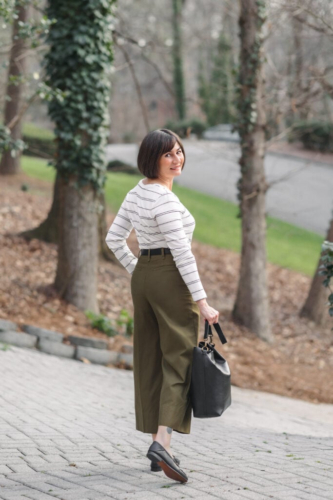 Back view of Kelly walking in neutral striped v-neck tee tucked into green high-rise wide-leg cropped twill pants with black Loraine bit loafers and black Madewell bucket tote — showing wide-leg silhouette and cropped hem from behind