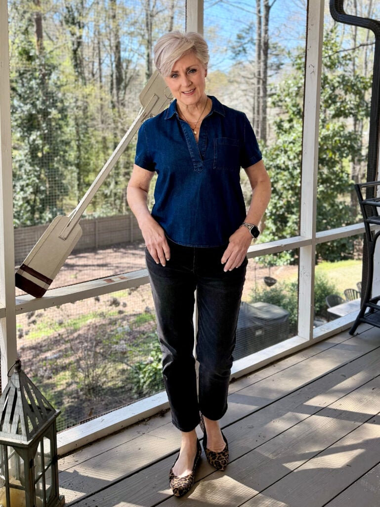 Beth stands on a screened porch in a dark denim short-sleeve polo top, black cropped jeans, and leopard-print pointed-toe flats, smiling at the camera with hands resting at her sides.