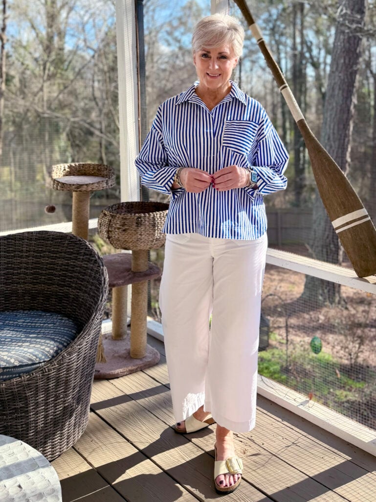 Full-length view of Beth in her striped popover and white wide leg crops, hands together, with Ollie's cat tree and a decorative oar visible on the porch