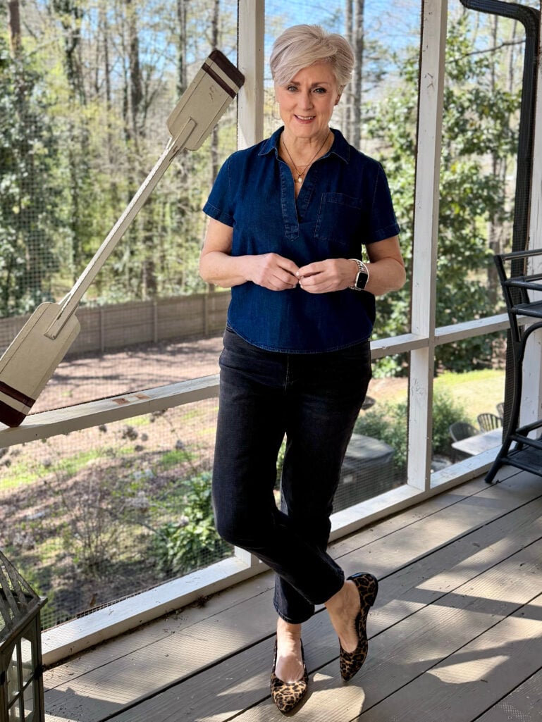 Beth leans casually against the screened porch railing in a dark denim polo top, black ankle jeans, and leopard flats, smiling with hands clasped in front of her.