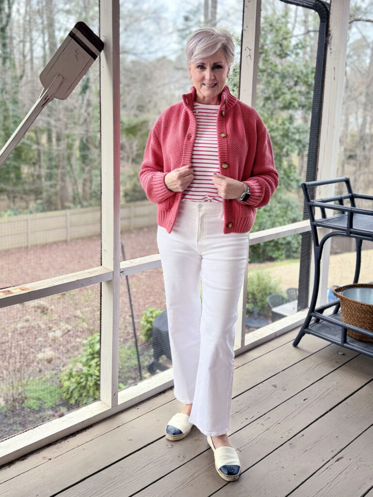 Full-length view of Beth in her coral sweater jacket and white denim, holding the jacket closed, with early spring greenery visible through the porch screens
