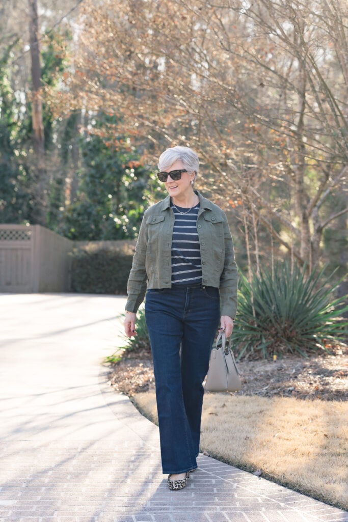 Classic style over 50 outfit with an olive utility jacket, striped top, dark jeans, and leopard flats.