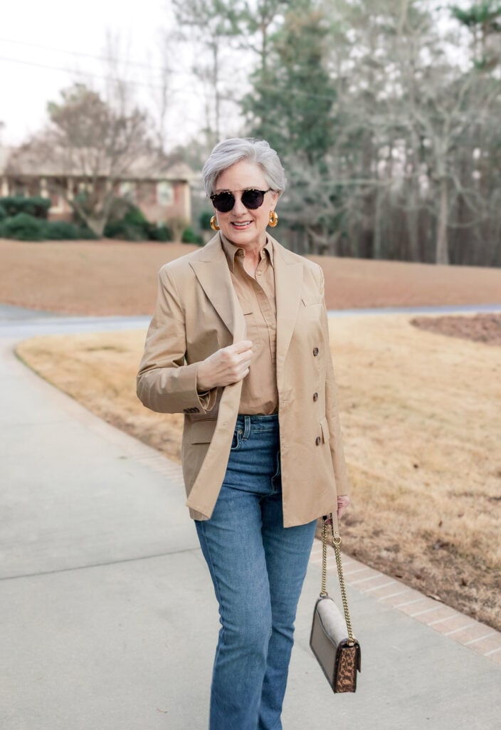 Woman over 50 with silver hair wearing a camel double-breasted blazer over a camel button-down shirt with medium-wash straight-leg jeans, gold hoop earrings, tortoiseshell sunglasses, and a taupe and leopard print chain-strap crossbody bag