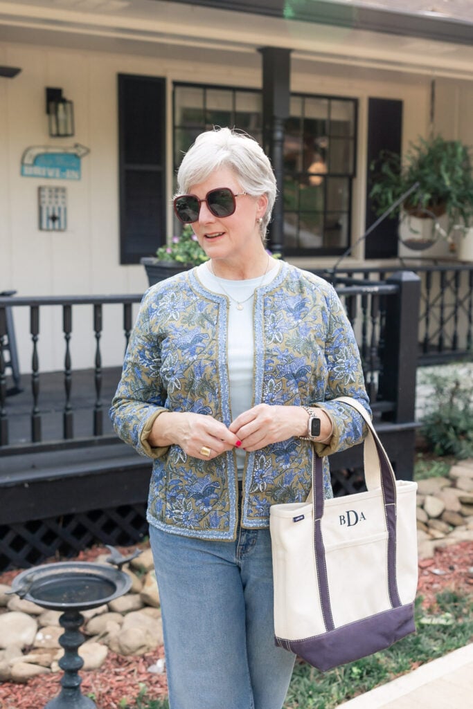 Woman over 50 with silver hair wearing blue and olive floral quilted jacket over white tee with medium wash jeans, carrying monogrammed canvas tote bag with navy trim, oversized sunglasses