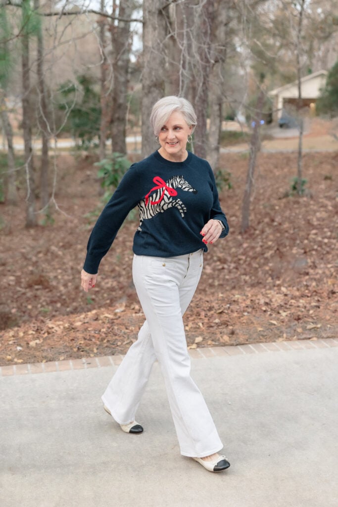 Woman over 50 with silver hair wearing navy crewneck sweater with zebra and red bow graphic, white flare jeans, and cream ballet flats with black cap toe