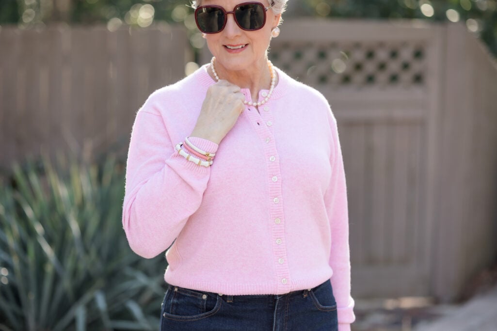 Close-up of woman over 50 with silver hair wearing soft pink button-front cardigan with pearl necklace, stacked pink and white bangles, and oversized sunglasses