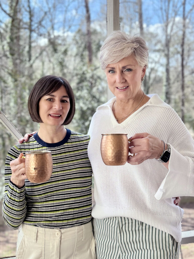 Kelly and Beth holding hammered copper mule mugs on the screened porch, Kelly in her green striped crewneck sweater and Beth in her white collared sweater