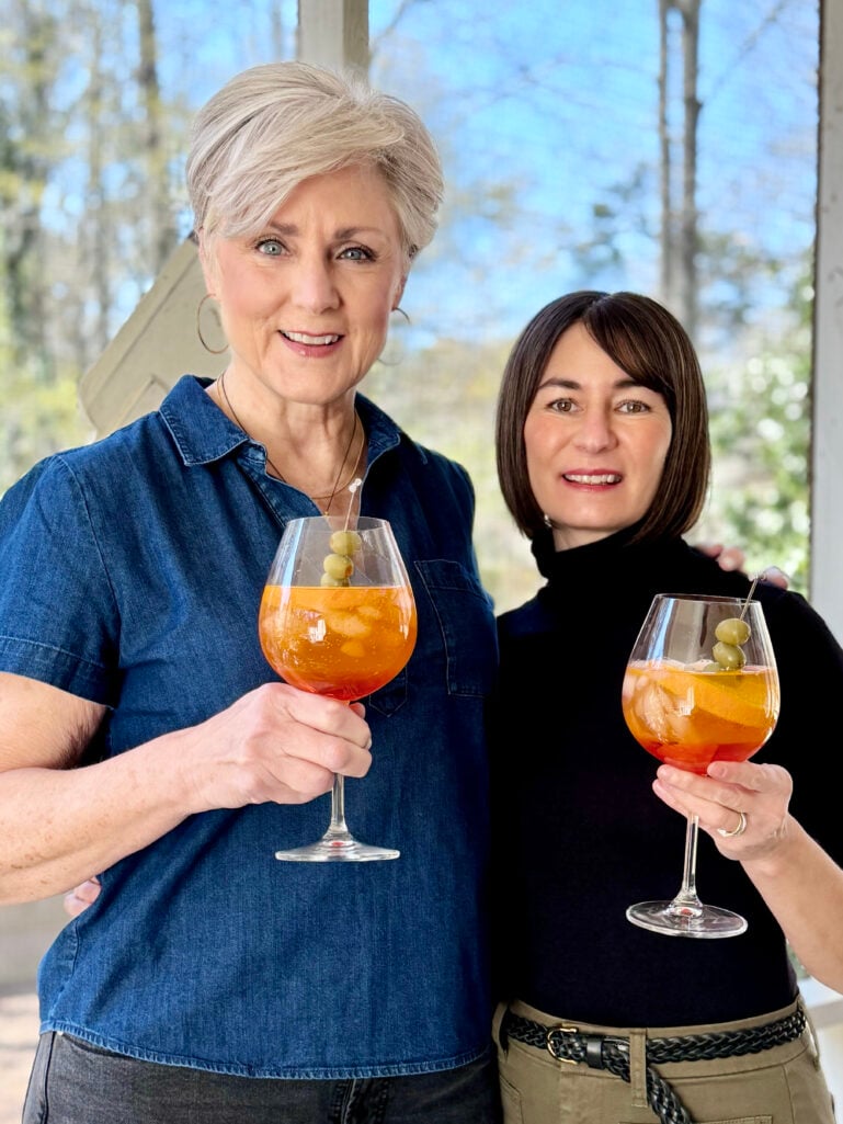 Beth and Kelly smile at the camera on a sunlit screened porch, each holding a stemmed wine glass filled with an orange Aperol Spritz garnished with olives and an orange slice.