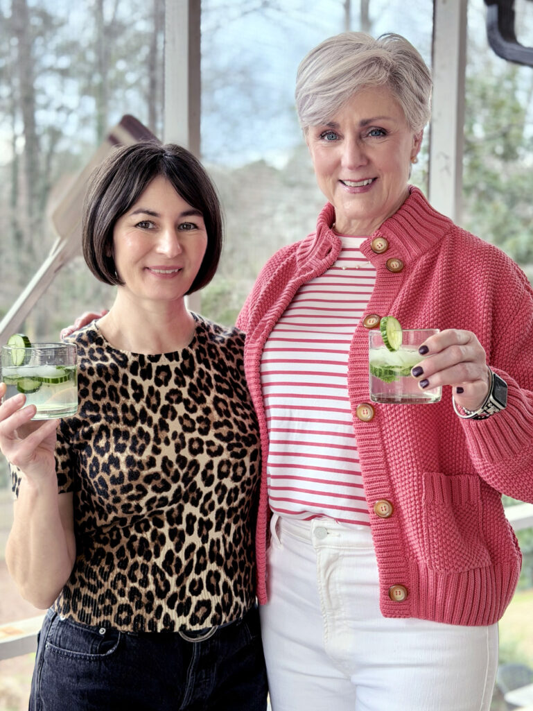 Kelly and Beth holding Cucumber Gin & Tonics in rocks glasses with cucumber wheels, Kelly in her leopard print sweater and Beth in her coral sweater jacket on the screened porch