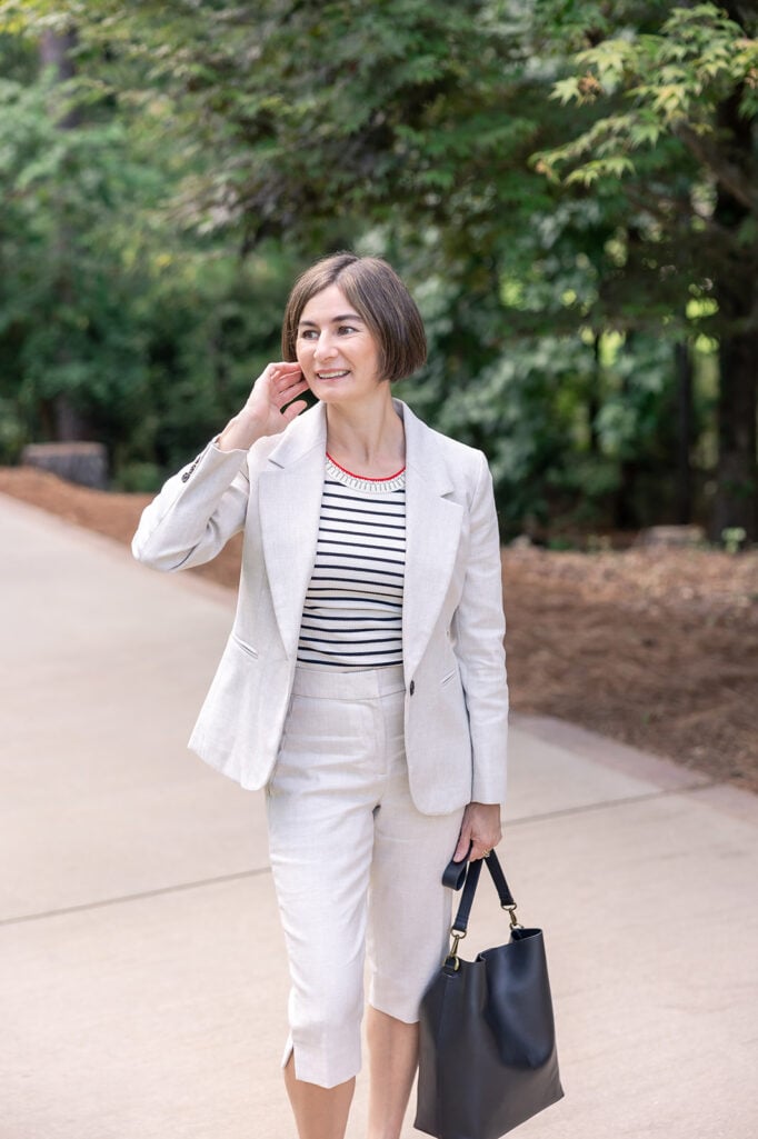Woman with dark bob haircut wearing a light gray linen blazer and matching bermuda shorts with a navy and white stripe tee and red beaded collar necklace, carrying a structured navy leather bucket bag with gold hardware — classic leather bucket bag spring 2026