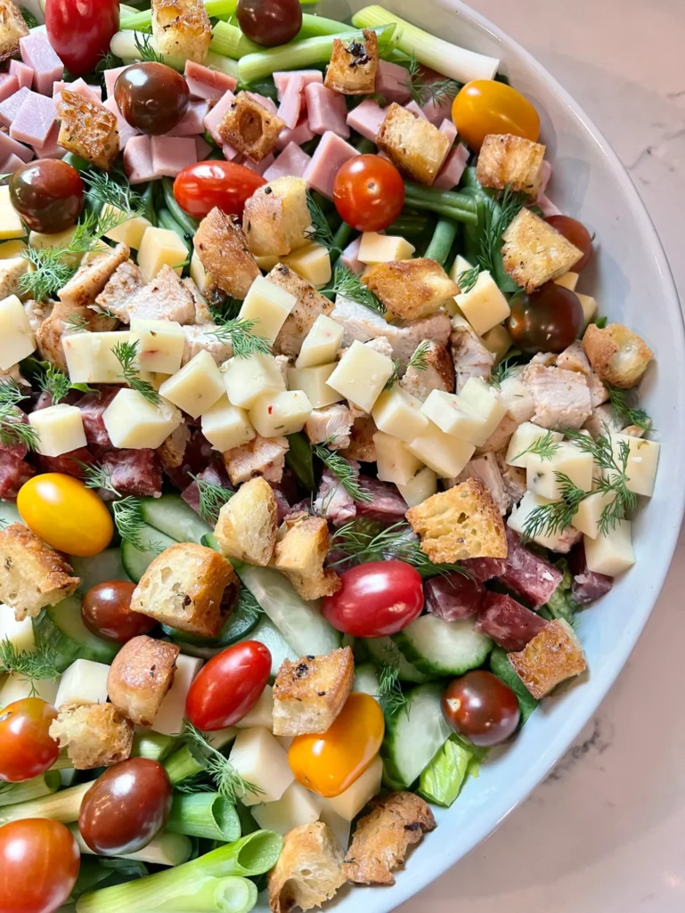 Close-up angle of chef salad showing diced chicken, cubed cheeses, salami, ham, croutons, cherry tomatoes, and sprigs of fresh dill