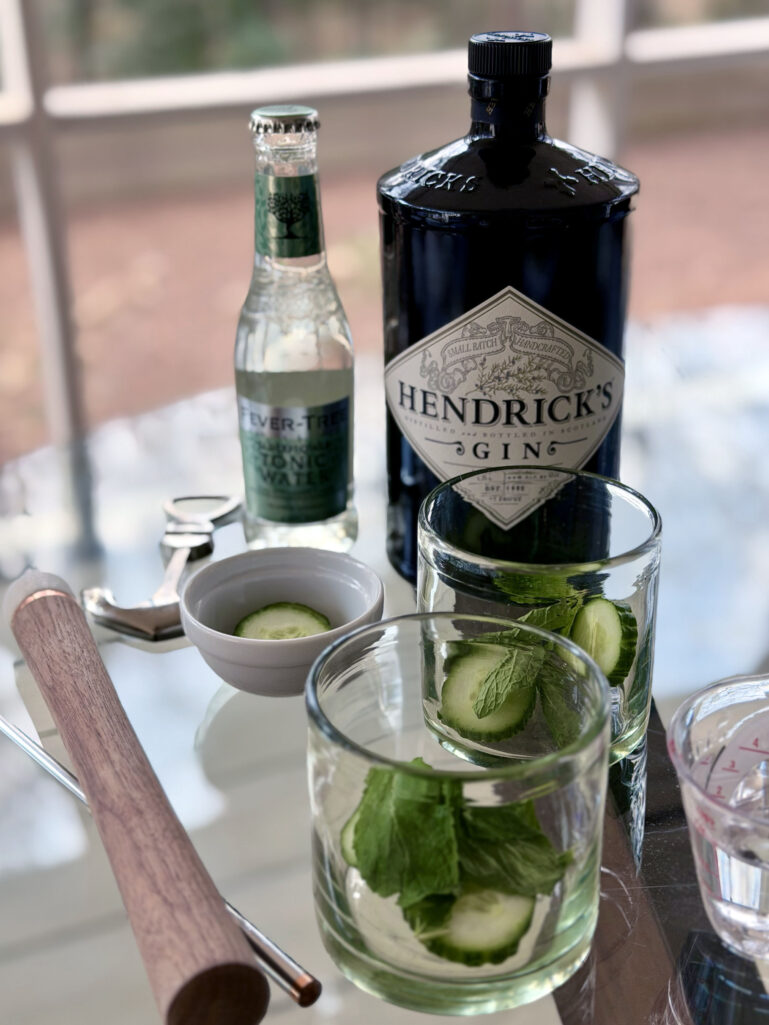 Another angle of the Cucumber Gin & Tonic setup with Hendrick's gin, Fever-Tree tonic, fresh cucumber and mint in a rocks glass, and a wooden muddler on the porch table