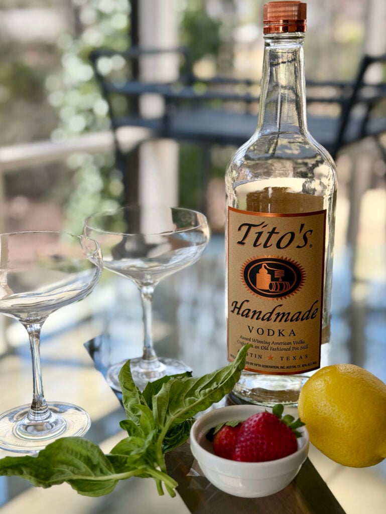 Strawberry Basil Smash ingredients on a glass-topped table: Tito's Handmade Vodka, fresh basil leaves, strawberries in a small white bowl, a whole lemon, and empty coupe glasses catching the afternoon light