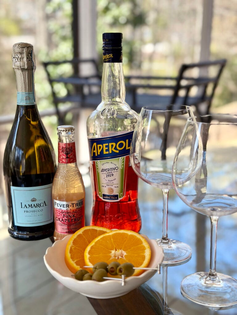 Aperol Spritz ingredients on a glass table: a bottle of La Marca Prosecco, a small bottle of Fever-Tree Sparkling Pink Grapefruit, a bottle of Aperol, two large empty wine glasses, and a small bowl of sliced oranges and green olives on cocktail picks.