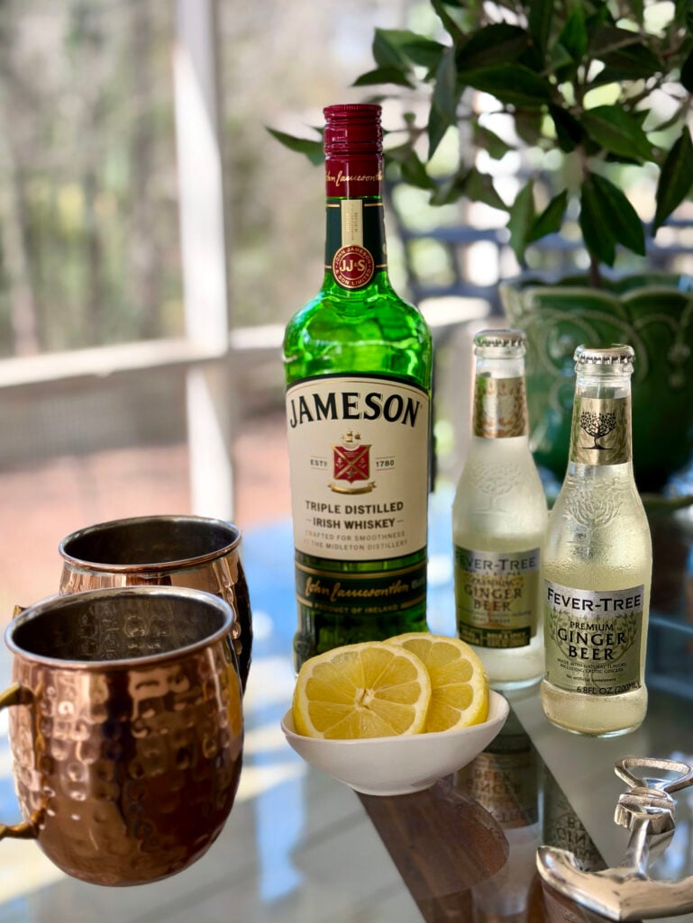 Irish Mule ingredients arranged on a glass-topped table: Jameson Irish Whiskey, two bottles of Fever-Tree Premium Ginger Beer, hammered copper mule mugs, and sliced lemons in a small white bowl