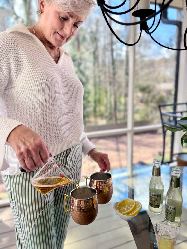 Beth pouring Jameson into hammered copper mule mugs from a glass measuring cup, Fever-Tree Ginger Beer bottles and lemon slices alongside, wearing her white sweater and green striped chinos