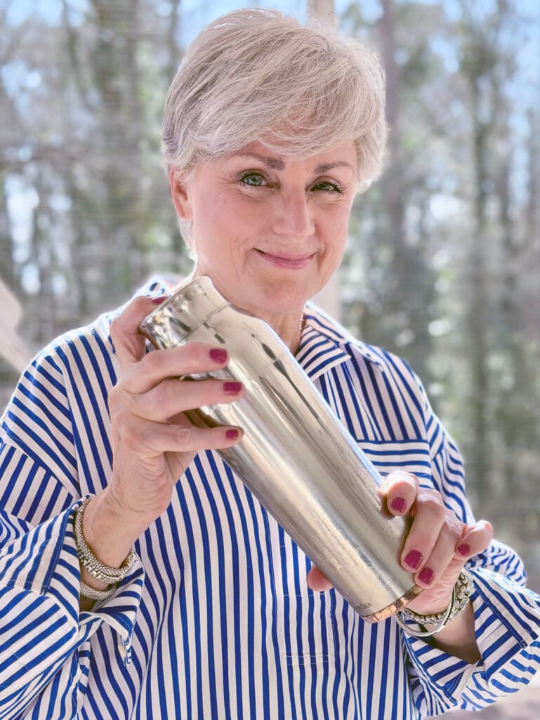 Close-up of Beth shaking a cocktail shaker with a playful smile, her blue and white striped popover sleeves billowing, spring trees soft in the background