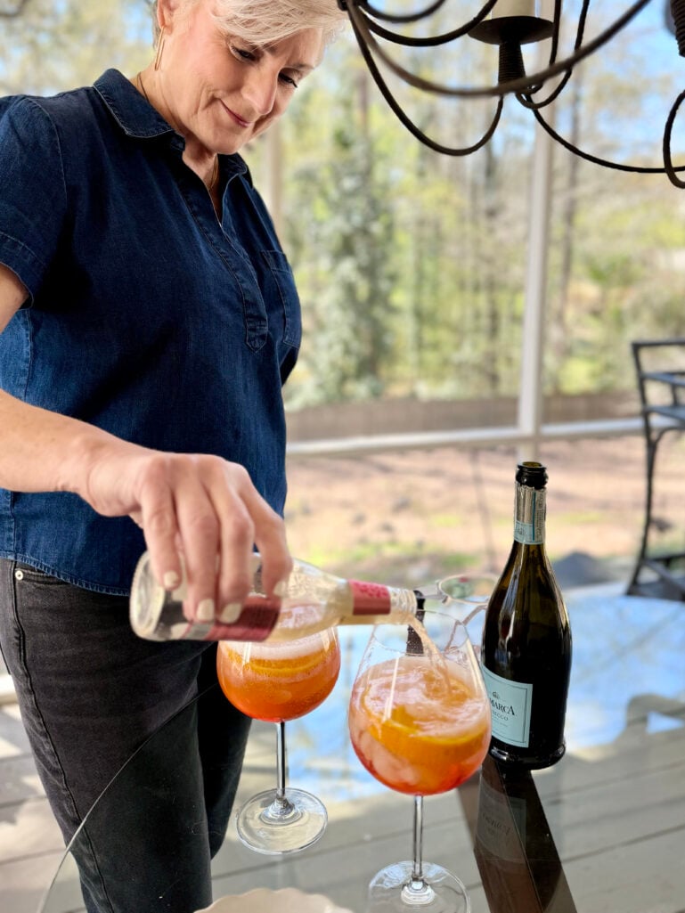 Beth pours Fever-Tree Sparkling Pink Grapefruit into two stemmed wine glasses already filled with Aperol and orange slices, with a bottle of La Marca Prosecco nearby on a glass table.