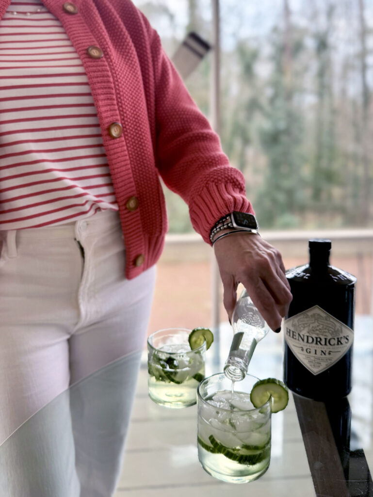 Close-up of Beth pouring Fever-Tree tonic water into rocks glasses with cucumber and mint over ice, her coral sweater jacket and watch visible, Hendrick's gin bottle alongside